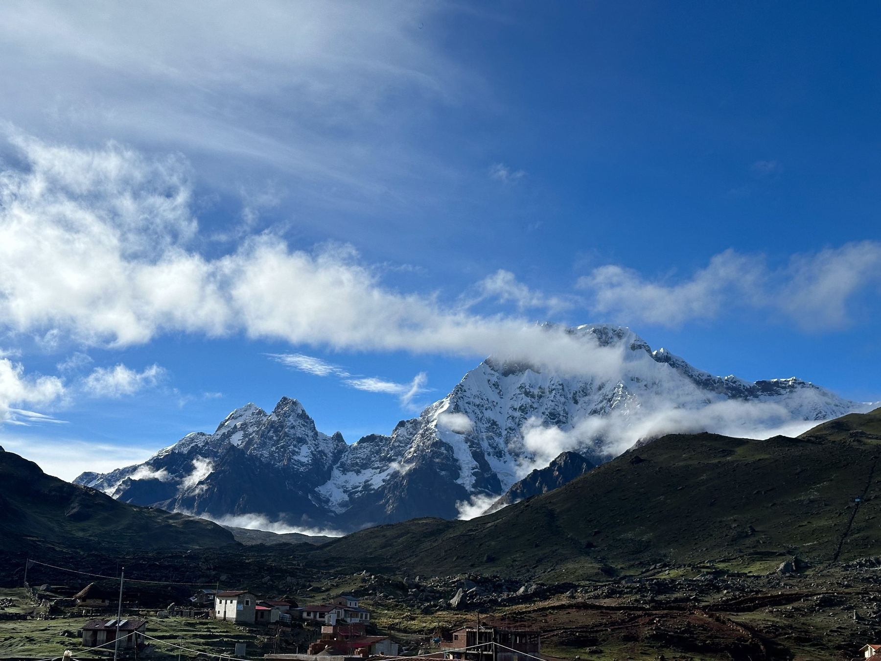 Snow-capped mountain peak above green hills and a small village, under a blue sky with clouds