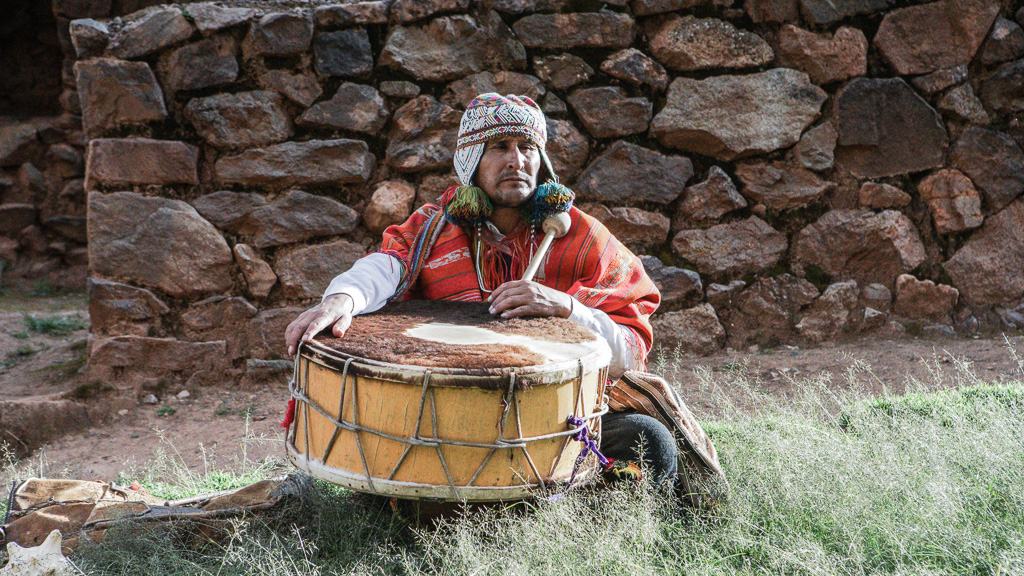 Man in a woolen cap and red poncho sitting behind a large drum, in front of a stone wall