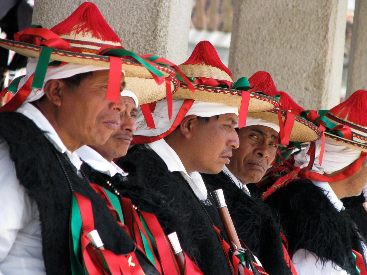 Men in black wool vests and wide straw hats with red tassels and green and red ribbons, seated in a row