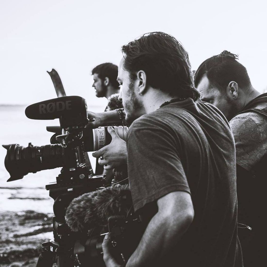Black and white photo of Iván looking into a film camera on a shore, with two colleagues behind him