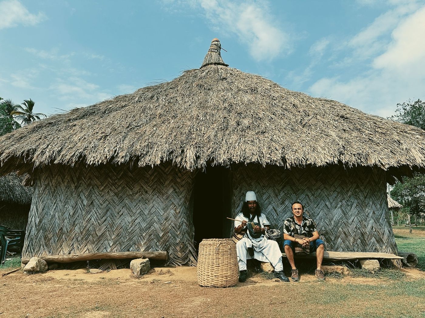 Iván sitting on a bench beside a man dressed in white, in front of a large thatched round house in the Sierra Nevada de Santa Marta